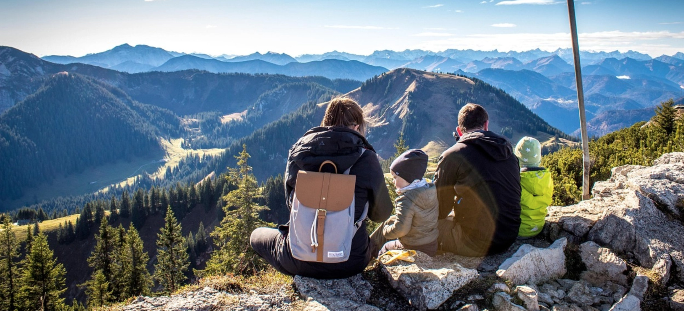 Eine Familie mit Wanderausrüstung ruht sich sitzend auf dem Gipfel eines Berges aus und geniesst die Aussicht ins Tal.