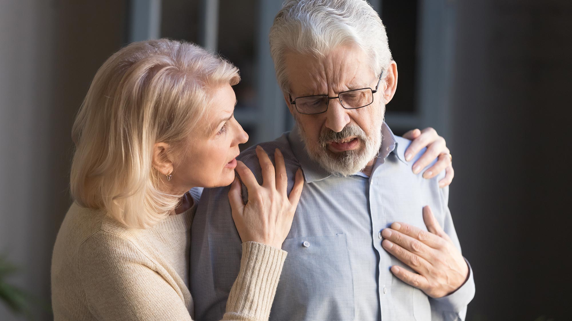 Ein älterer Mann legt besorgt seine Hand auf die Brust. Eine ebenfalls besorgte Frau steht daneben und nimmt ihn in den Arm.