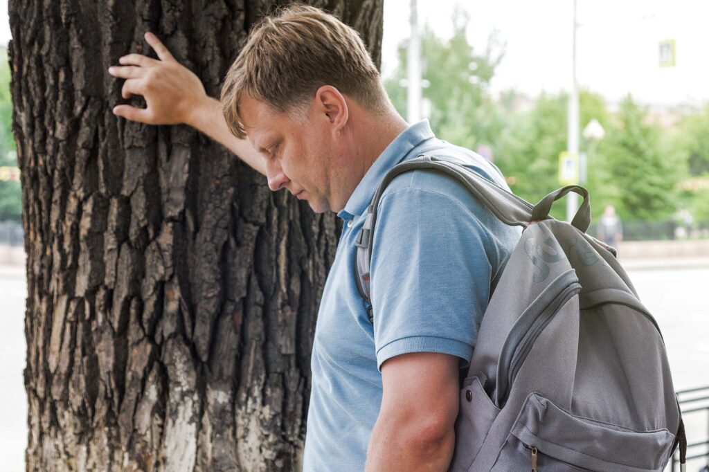 Ein Mann mittleren Alters mit einem Rucksack lehnt sich an einen Baumstamm.