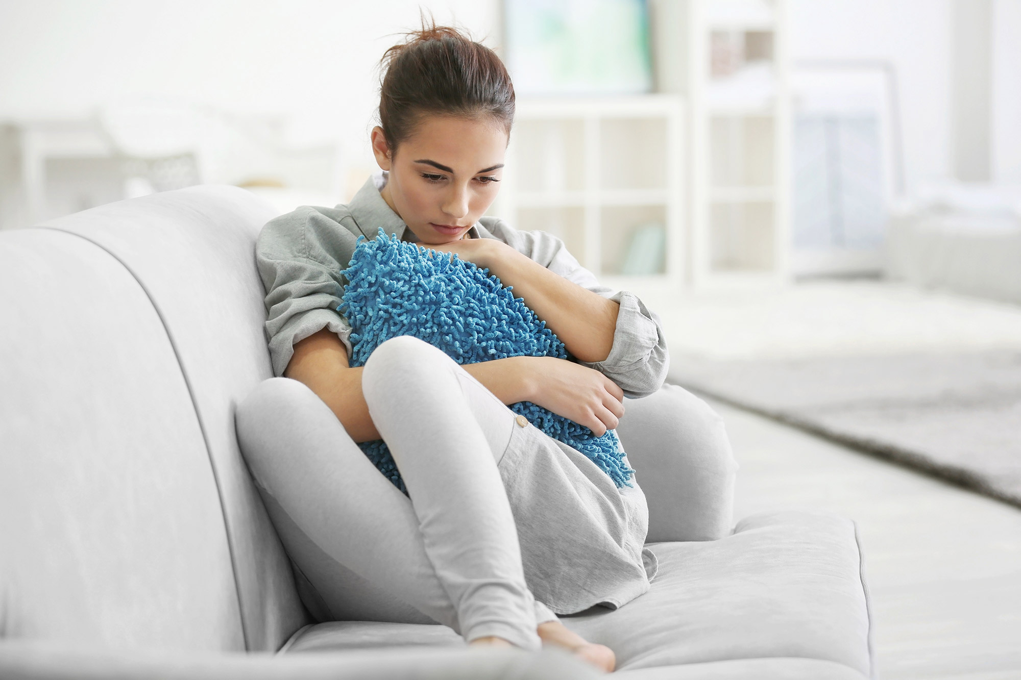 Eine junge Frau sitzt nachdenklich mit angewinkelten Beinen auf einem Sofa und umklammert ein Kissen.