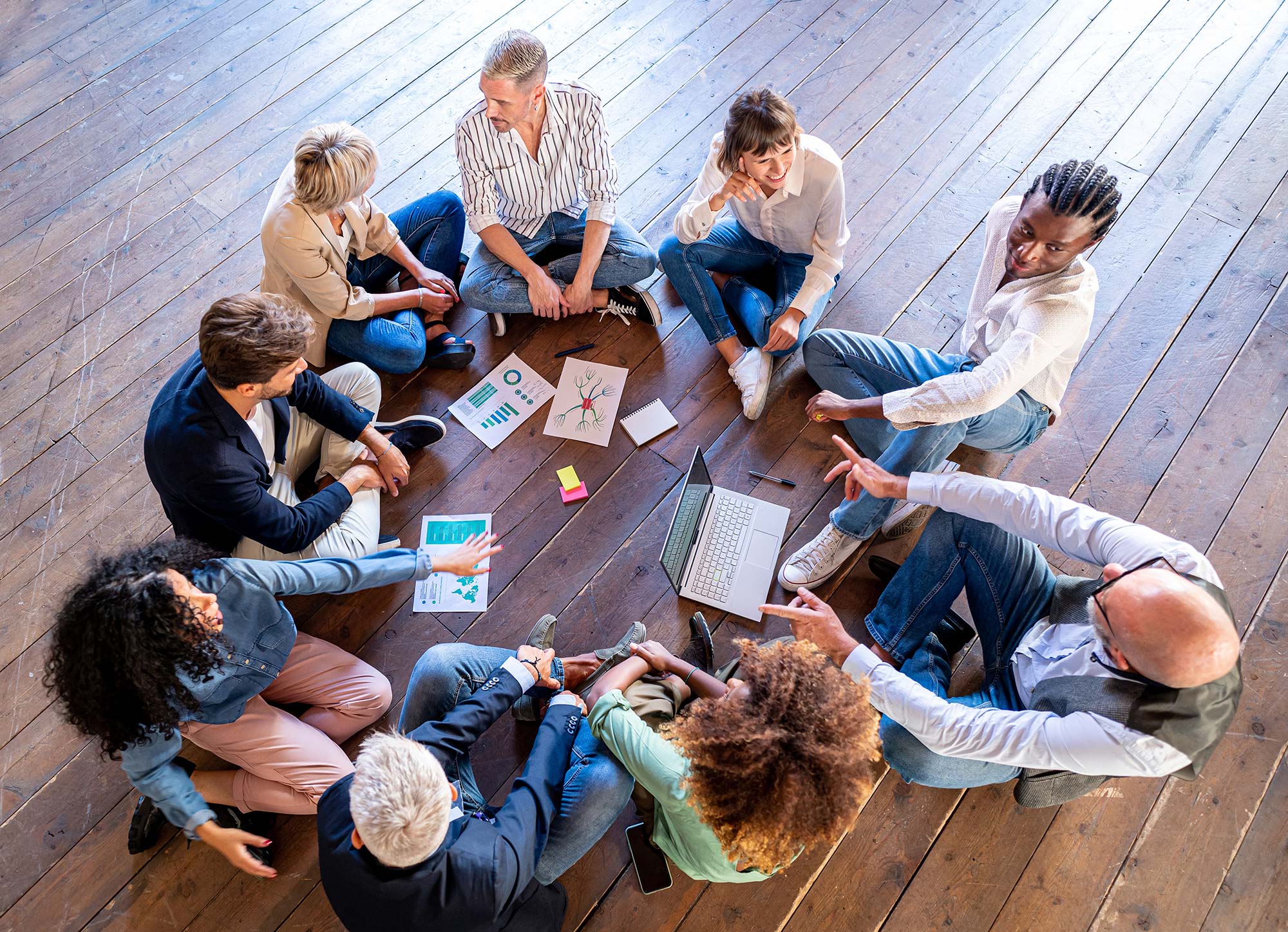 Neun Personen sitzen auf einem Holzboden im Kreis und halten eine Besprechung ab.
